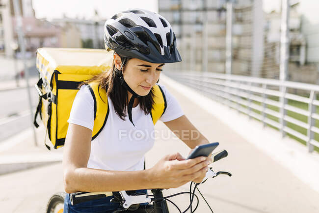 Mulher de entrega verificando o telefone celular enquanto está de pé com bicicleta na estrada — Fotografia de Stock