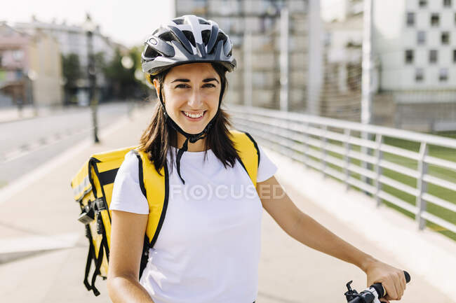 Sorrindo mulher serviço essencial vestindo capacete de ciclismo e mochila de entrega no dia ensolarado — Fotografia de Stock