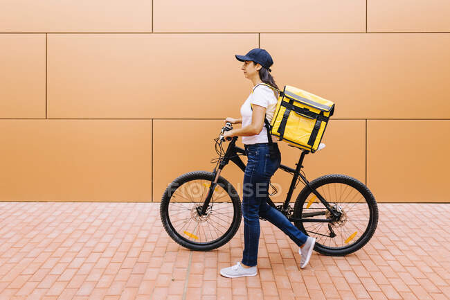 Delivery woman with backpack wheeling bicycle on footpath — Stock Photo