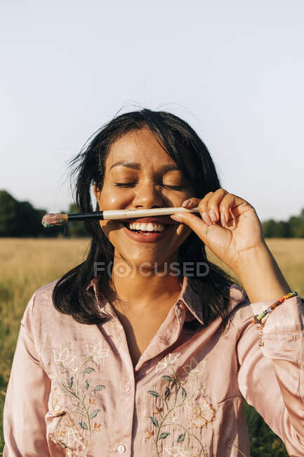 Mujer sonriente con los ojos cerrados sosteniendo pincel durante el día soleado - foto de stock