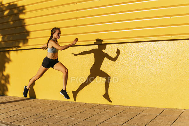 Sportswoman running by yellow wall during sunny day — Stock Photo