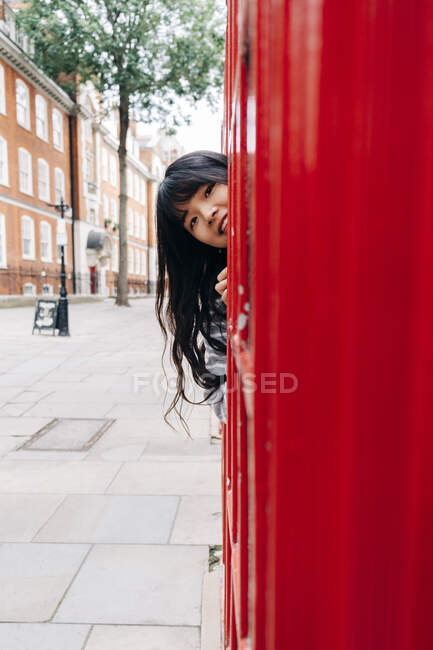 Playful woman peeking by telephone booth in city — Stock Photo