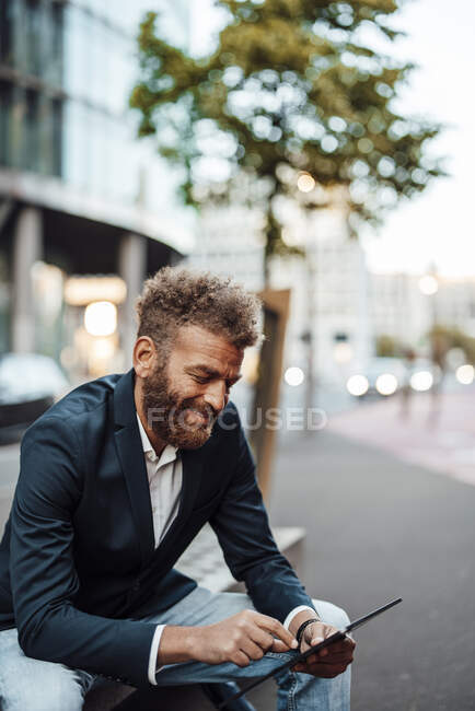 Freelancer masculino sonriente trabajando en una tableta digital mientras está sentado en el banco - foto de stock