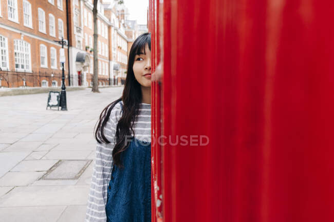 Young woman with long hair standing behind telephone booth in city — Stock Photo
