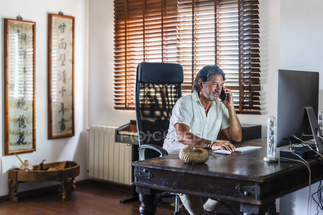 Hombre profesional que utiliza la computadora mientras habla en el teléfono inteligente en casa - foto de stock