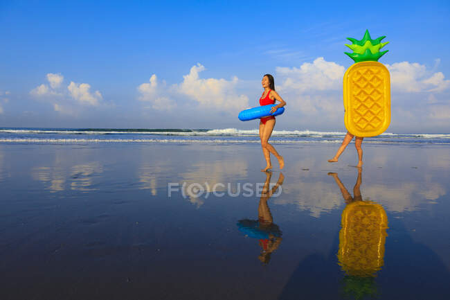 Mujeres amigas sosteniendo flotadores nadando mientras caminan en la playa - foto de stock