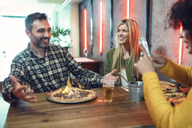 Jovem mulher fotografando homem gesticulando na frente de pizza flamejante por amigo feminino no restaurante — Fotografia de Stock