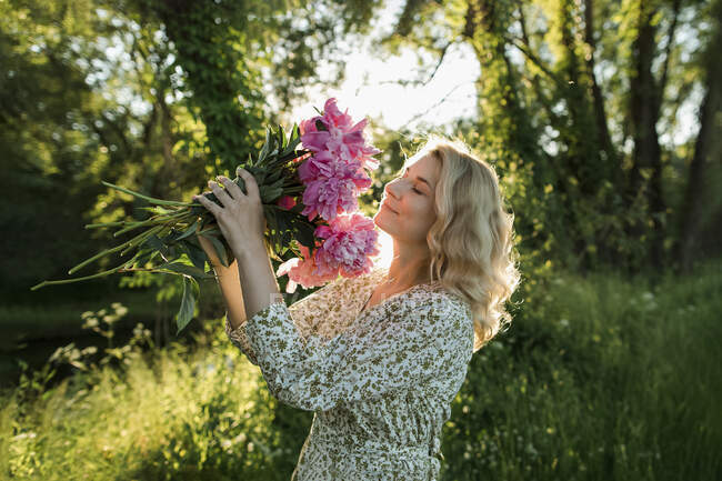 Blonde Frau riecht Pfingstrosenblume, während sie auf der Wiese steht — Stockfoto