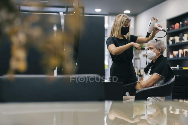 Friseurin mit Gesichtsmaske föhnt Kundin im Salon die Haare — Stockfoto