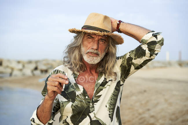 Hombre ajustando sombrero mientras sostiene el cigarrillo en la playa - foto de stock