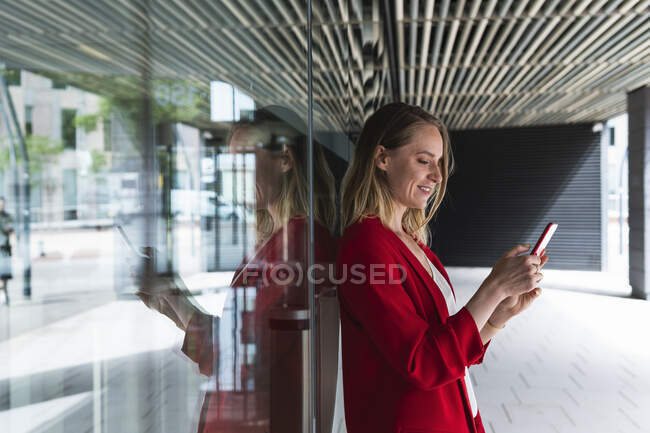 Femme professionnelle utilisant un téléphone portable tout en s'appuyant sur le mur de verre — Photo de stock