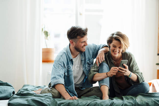 Hombre sentado con la mujer usando el teléfono inteligente en la cama en casa - foto de stock