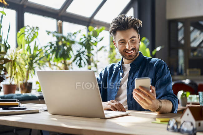 Усміхнений чоловік-професіонал використовує мобільний телефон, сидячи за столом в офісі — стокове фото