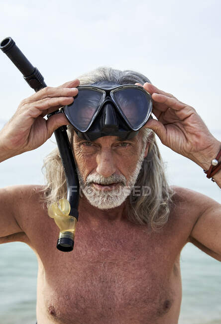 Hombre maduro sin camisa sosteniendo gafas de snorkel en la playa - foto de stock