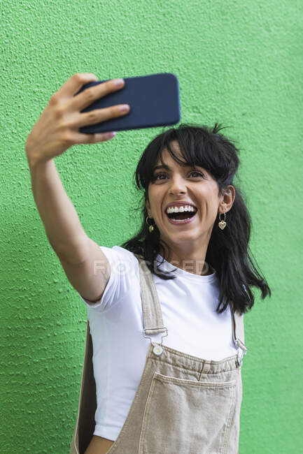 Femme gaie prenant selfie par téléphone intelligent contre le mur vert — Photo de stock