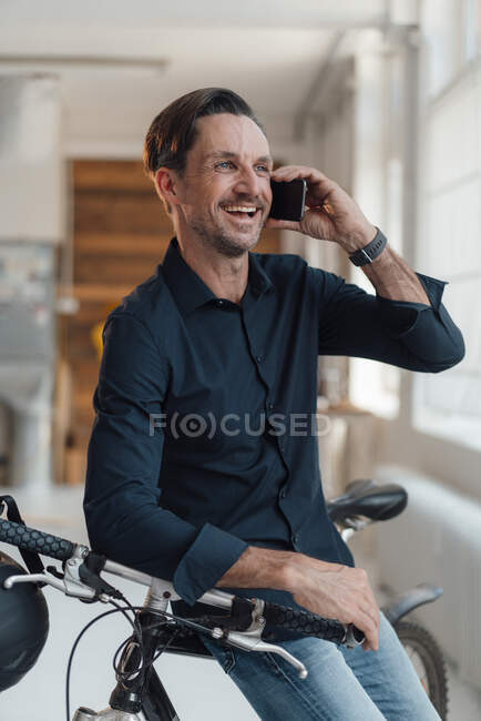 Feliz hombre de negocios maduro hablando por teléfono inteligente mientras está sentado en bicicleta en la oficina - foto de stock