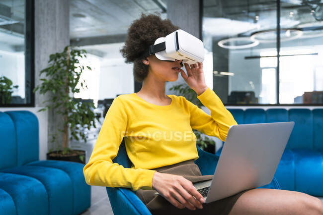 Femme d'affaires avec ordinateur portable portant casque de réalité virtuelle tout en étant assis dans le hall du bureau — Photo de stock