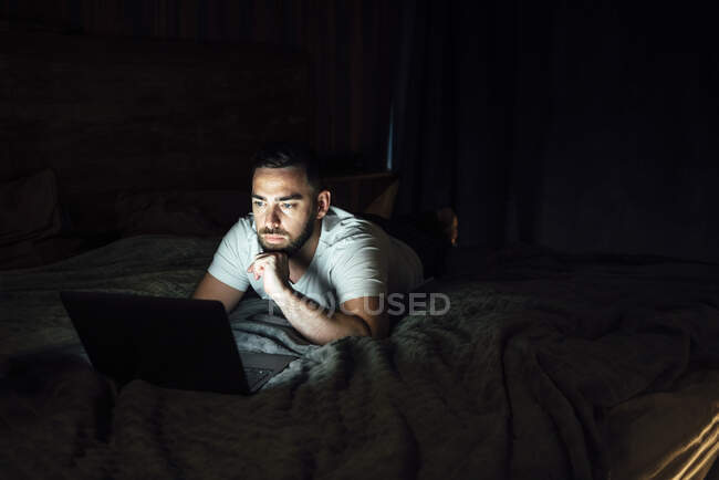 Задумчивый человек использует ноутбук, лежа на кровати дома — стоковое фото