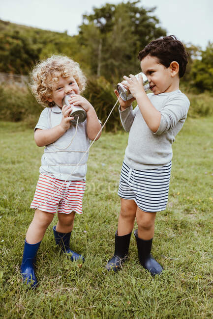 Felices amigos masculinos hablando a través de lata puede telefonear mientras juega en el prado - foto de stock