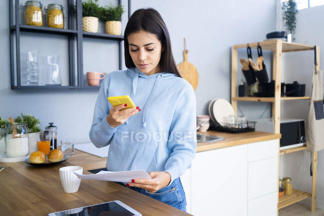 Mujer fotografiando documento a través del teléfono móvil en el mostrador de cocina - foto de stock