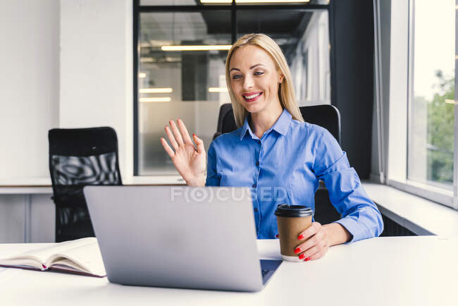 Smiling blond businesswoman waving hand during video call through laptop in office — Stock Photo