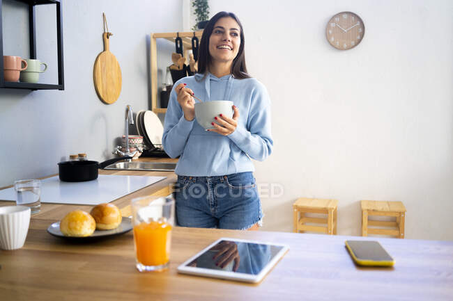 Улыбающаяся молодая женщина держит миску у кухонного стола дома — стоковое фото