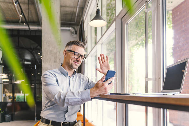 Чоловік, який махає рукою під час відеодзвінка через мобільний телефон у офісі. — стокове фото