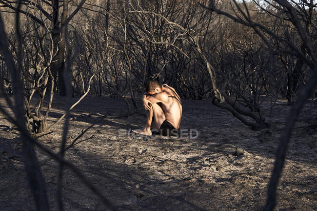 Hombre sentado en posición fetal en el suelo del bosque quemado - foto de stock