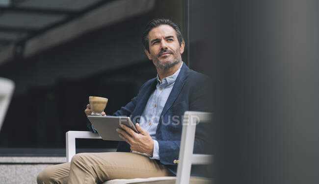 Зрілий чоловік тримає цифрову табличку і чашку кави, сидячи на стільці. — стокове фото