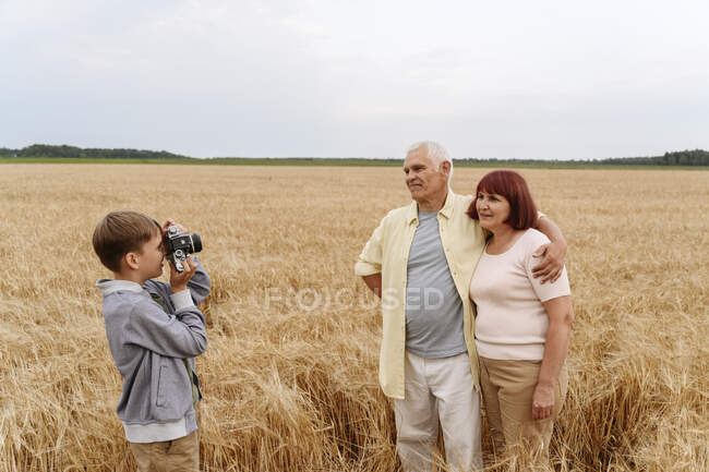 Petit-fils photographier les grands-parents à travers la caméra dans le champ de blé — Photo de stock