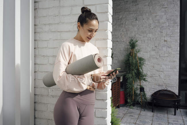 Femme moyenne adulte avec tapis de yoga en utilisant le téléphone mobile sur la terrasse — Photo de stock