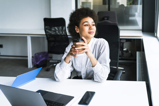 Mujer de negocios sosteniendo taza de café desechable mientras está sentado con el ordenador portátil en la oficina - foto de stock