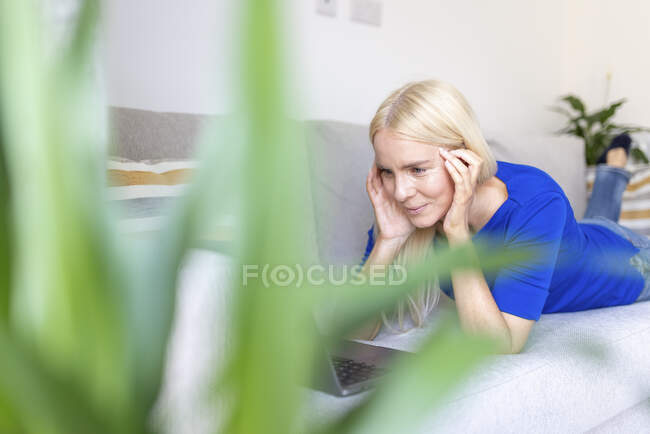 Donna con la testa in mano guardando il computer portatile mentre giaceva sul divano a casa — Foto stock