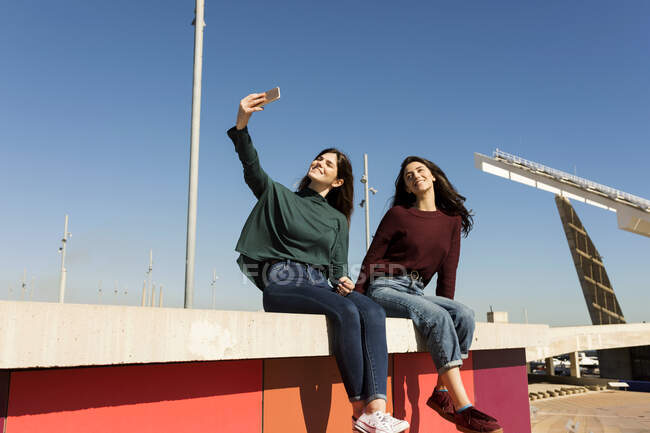 Женщина делает селфи, сидя с подругой на стене — стоковое фото