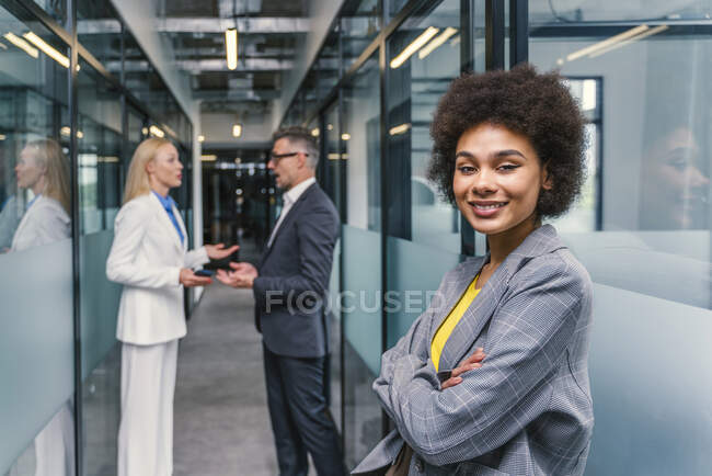 Joven empresaria de pie con los brazos cruzados mientras hombre y mujer colega discutiendo en el pasillo - foto de stock