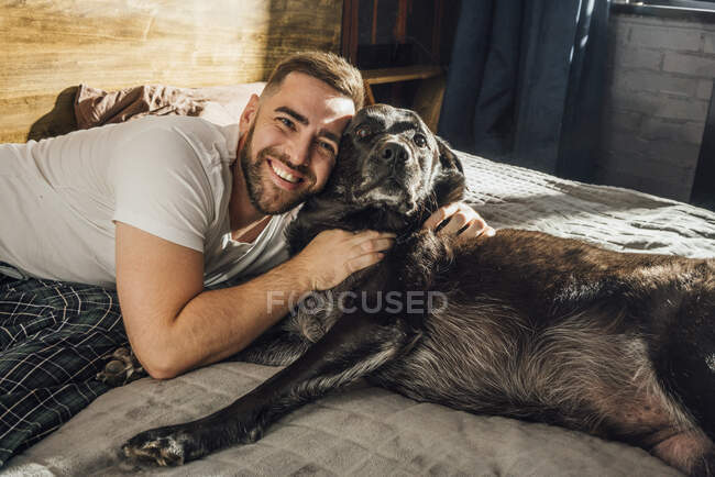 Sonriente hombre guapo abrazando perro en la cama en casa - foto de stock