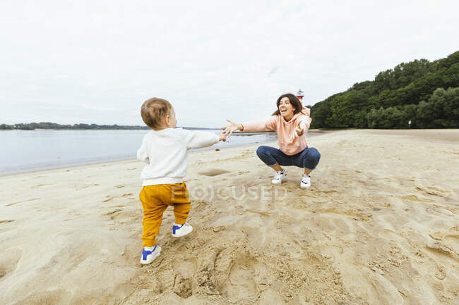 Male toddler walking towards mother crouching on sand at beach — Stock Photo