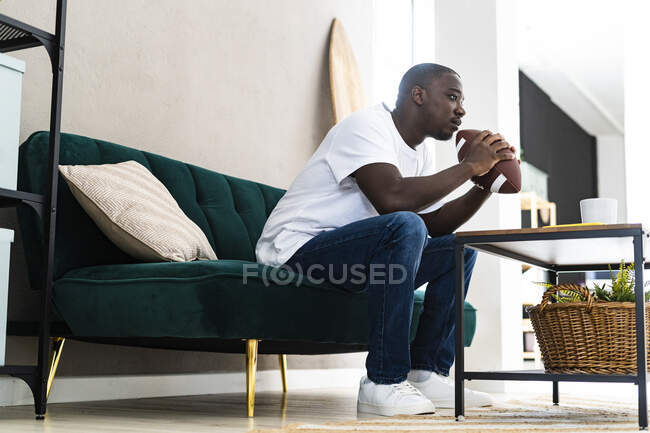Homme mûr réfléchi avec ballon de rugby assis sur le canapé à la maison — Photo de stock