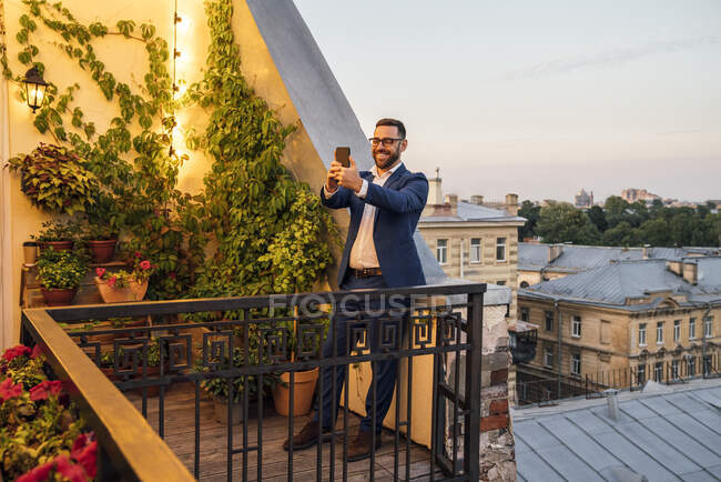 Улыбающийся бизнесмен делает селфи через смартфон на балконе во время заката — стоковое фото