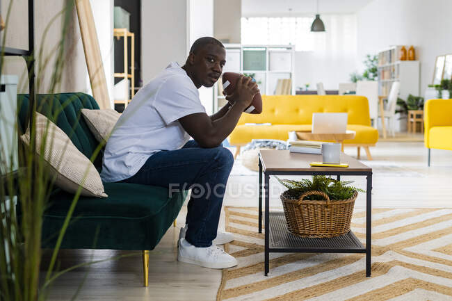 Hombre sosteniendo la pelota de rugby mientras está sentado en el sofá en casa - foto de stock