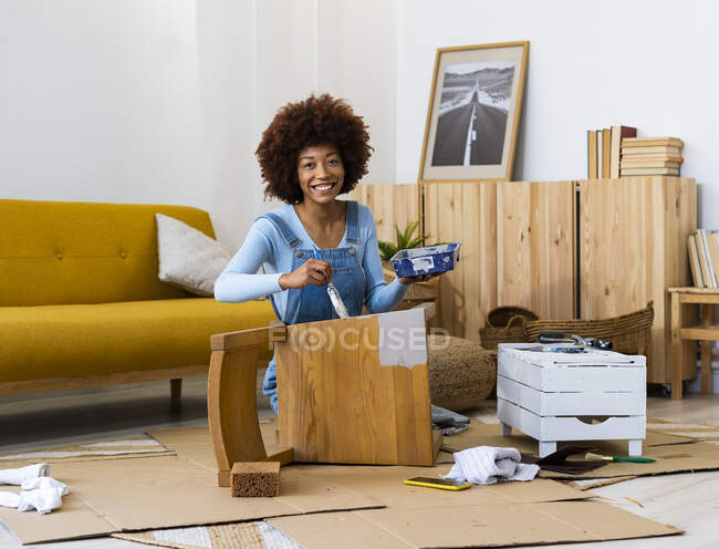 Happy young woman with paint tray painting old chair in living room — Stock Photo