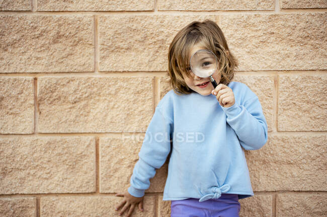 Cute girl through magnifying glass while standing in front of wall — Stock Photo