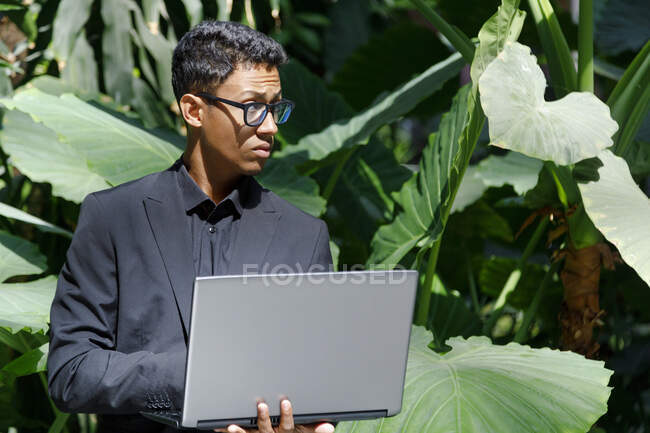 Jeune professionnel masculin avec ordinateur portable debout devant les plantes pendant la journée ensoleillée — Photo de stock