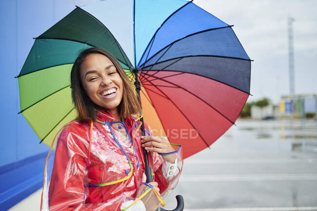 Mulher feliz vestindo capa de chuva segurando guarda-chuva multi colorido durante a estação chuvosa — Fotografia de Stock