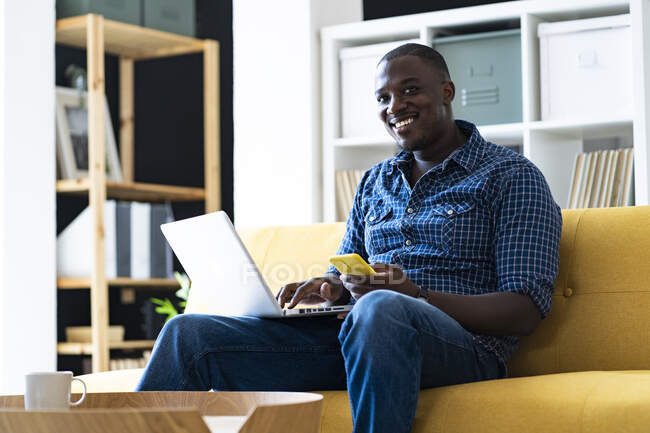 Чоловік з ноутбуком і смартфоном сидить на дивані вдома — стокове фото