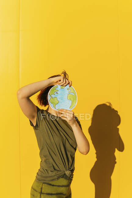 Mujer sosteniendo papel tierra planeta delante de la pared amarilla - foto de stock