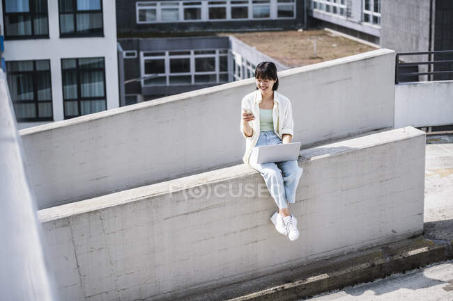 Smiling teenage girl with laptop using mobile phone while sitting on retaining wall — Stock Photo