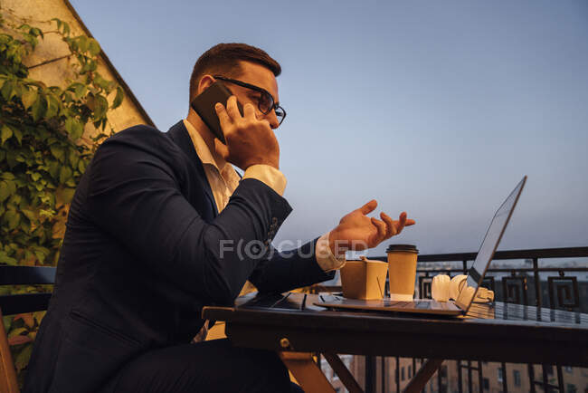 Uomo d'affari con computer portatile che parla su smartphone in balcone durante il tramonto — Foto stock