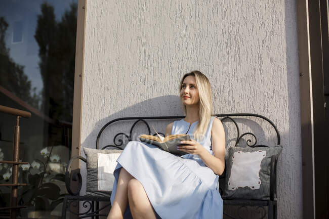 Nachdenkliche Frau mit Buch sitzt bei sonnigem Wetter auf Bank auf der Terrasse — Stockfoto