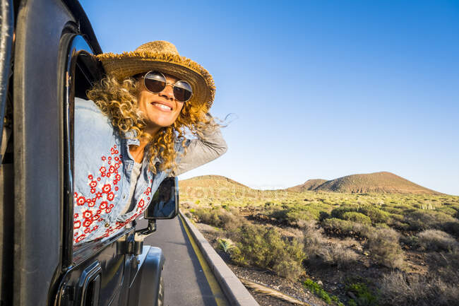 Mulher sorrindo usando chapéu inclinado para fora da janela do carro — Fotografia de Stock